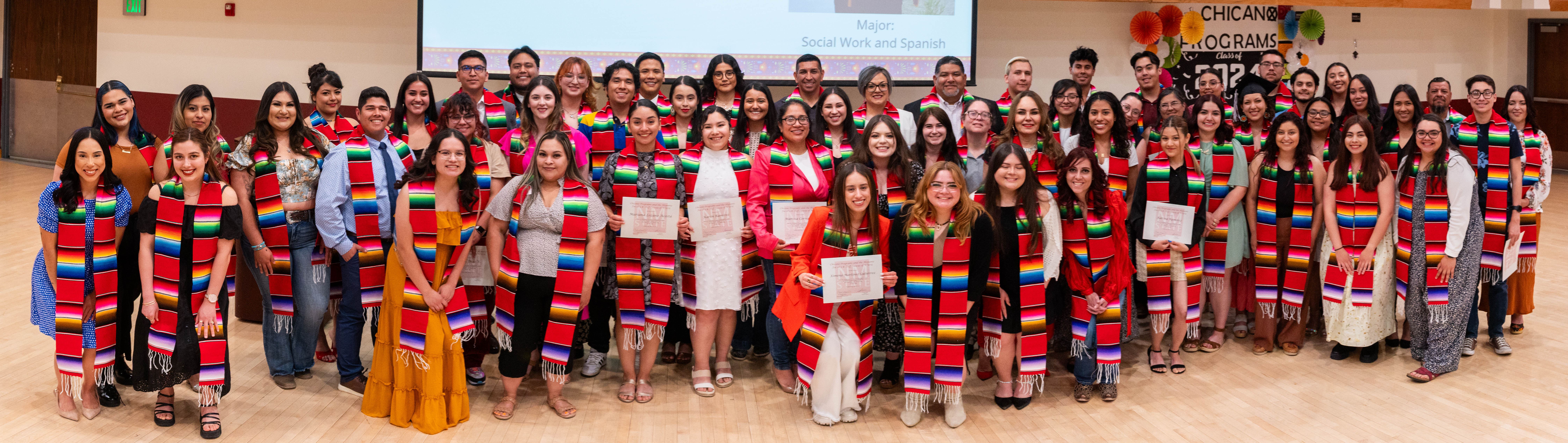 A photograph of all the students who attended Latinx Graduation. The image depicts a large group of people, predominantly young adults, standing side by side in a spacious, well-lit indoor area with a light wooden floor and beige walls. The attendees are wearing vibrant, multicolored serape stoles, which feature horizontal stripes in various bright colors, including red, blue, green, yellow, and white with fringed edges. Many are dressed in formal or semi-formal attire. Several of them hold certificates. In the background, there is a projected presentation on a screen with partially visible text, and various decorations including an arrangement reading "CHICANO PROGRAMS" and "Class of 2025."